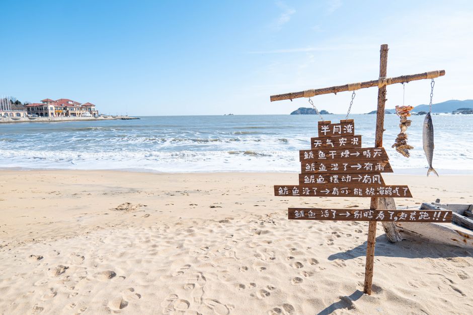 Wooden sign with fish on a sandy beach with ocean waves and distant architecture, Huizhou, Guangdong