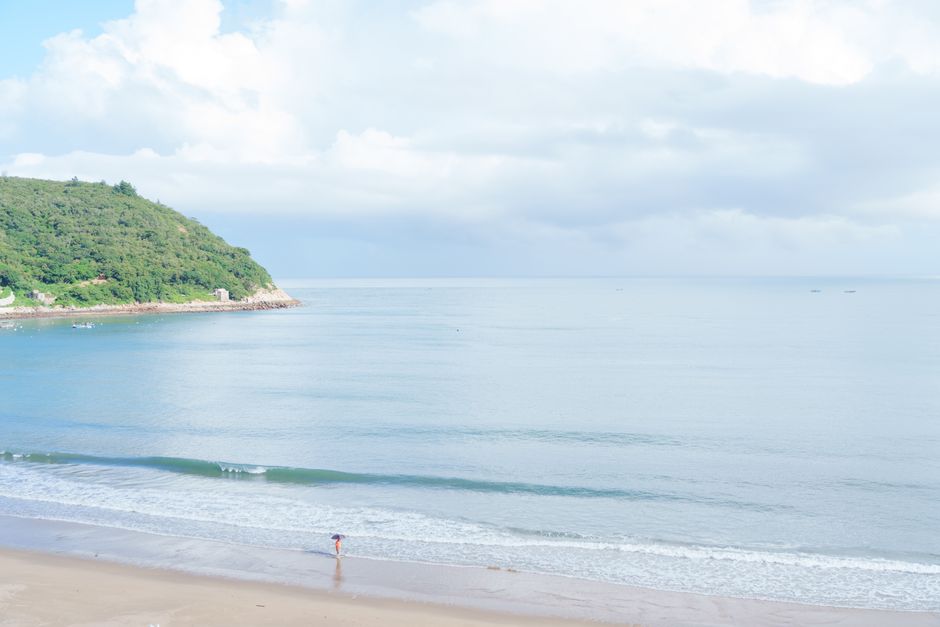 Peaceful beach scene with calm waters and green hill under blue sky, ideal for relaxation, Huizhou, Guangdong